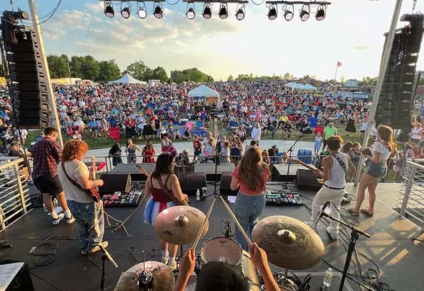 Widok z perspektywy sceny na festiwal muzyczny z tłumem ludzi i zespołem na scenie.